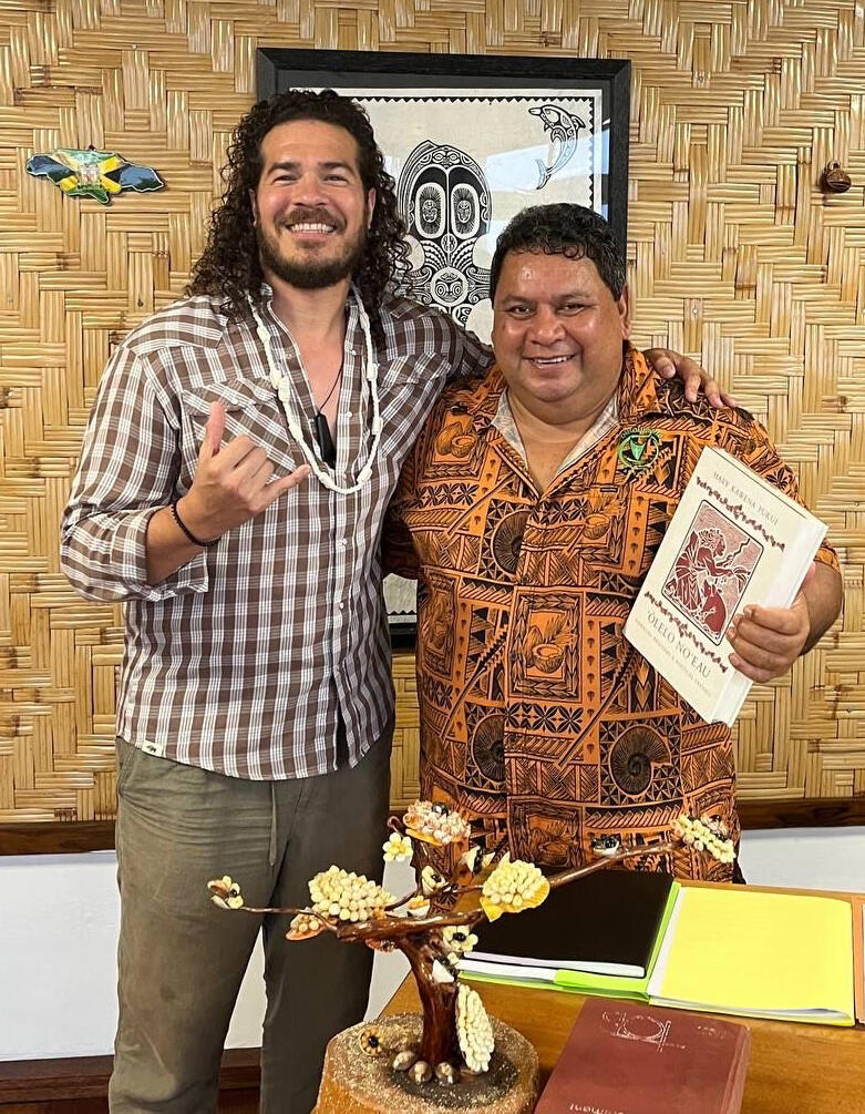 Two men in Polynesian shirts hugging, one making the Love-You hand gesture.